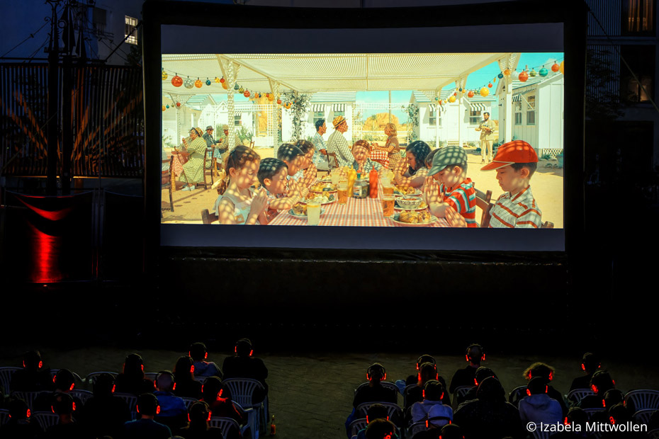 Es ist dunkel draußen. Viele Zuschauer mit Kopfhörern schauen zu einer Leinwand, wo ein Film mit mehreren Kindern betend an einem Tisch zu sehen ist.